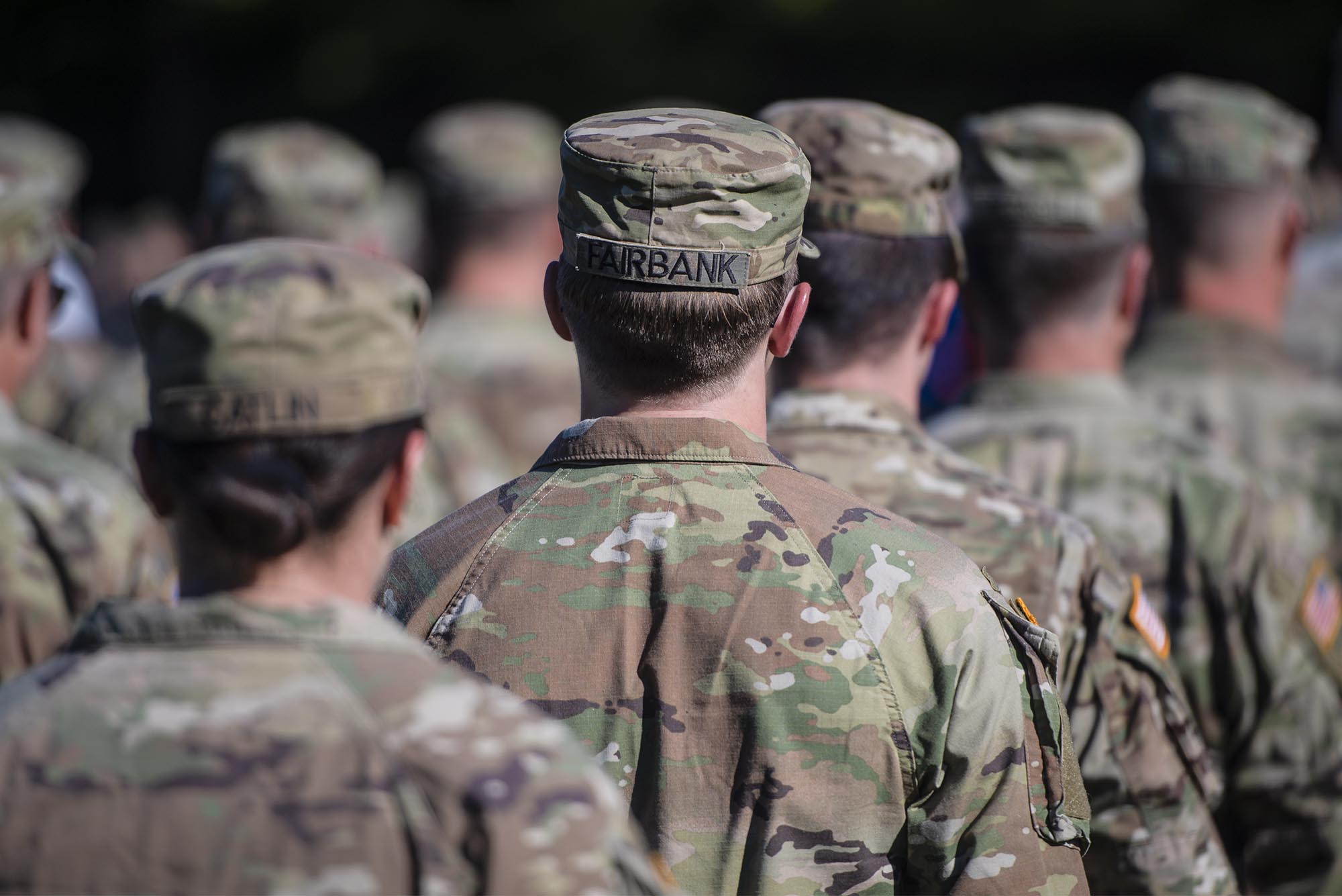 A group of soldiers in camouflage uniforms stand in formation, seen from behind, with names visible on the back of their caps.