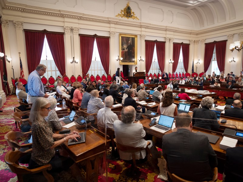 A government meeting in a grand room with high ceilings, red curtains, and numerous attendees seated and taking notes or using laptops. A large portrait hangs on the wall.