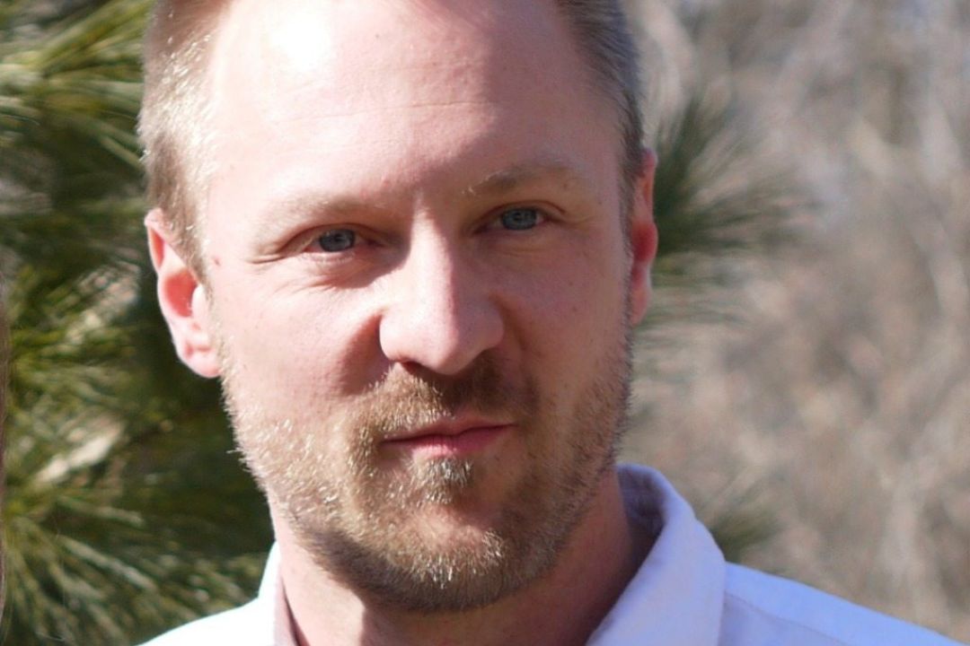 A man with short, light-colored hair and a beard, wearing a white shirt, stands outside with greenery in the background.