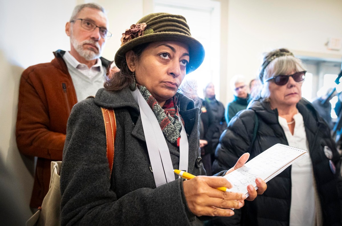 A woman in a hat takes notes with a pen and notebook in a room with other people.