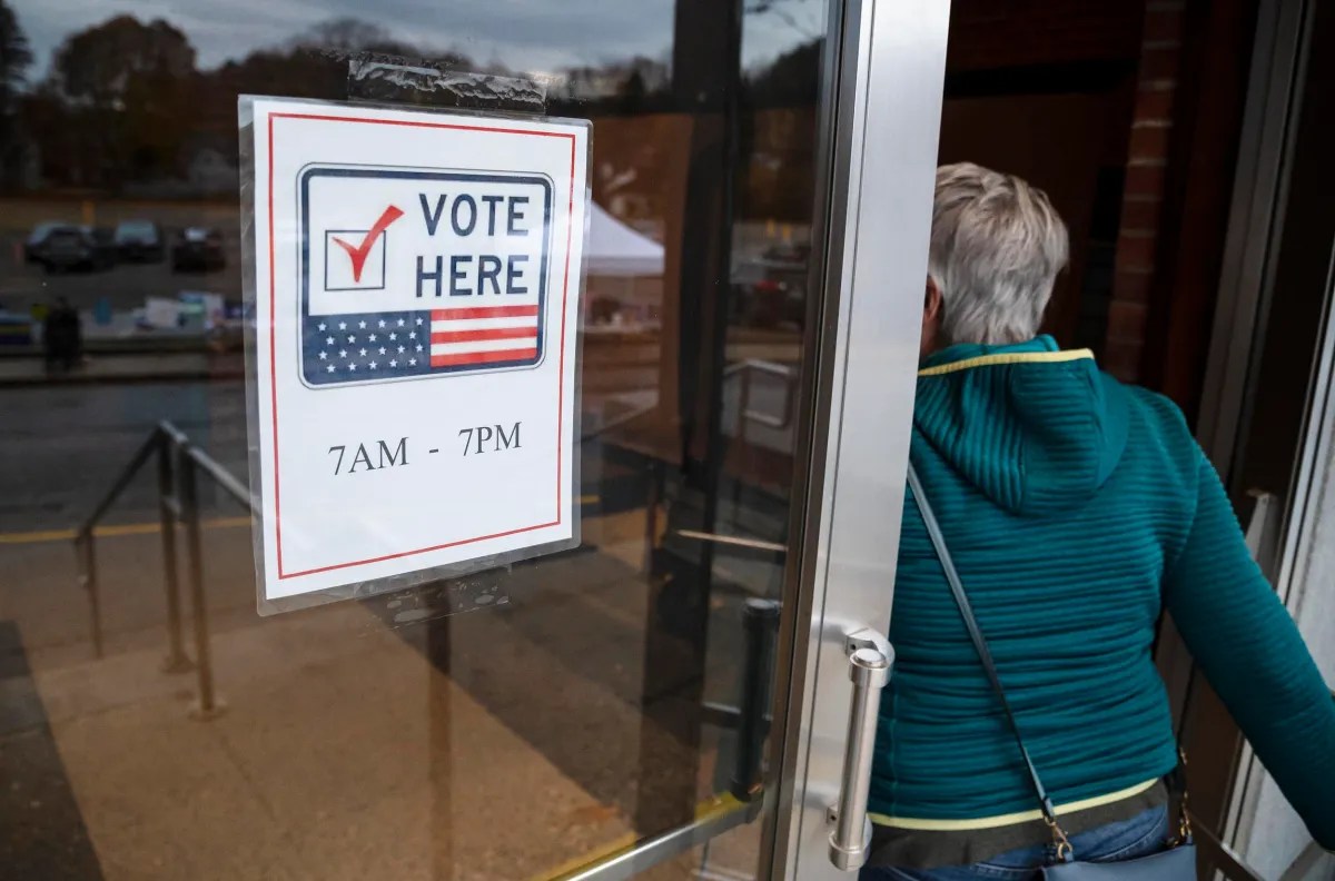 Person entering a polling station with a "Vote Here" sign on the door, indicating voting hours from 7 AM to 7 PM.