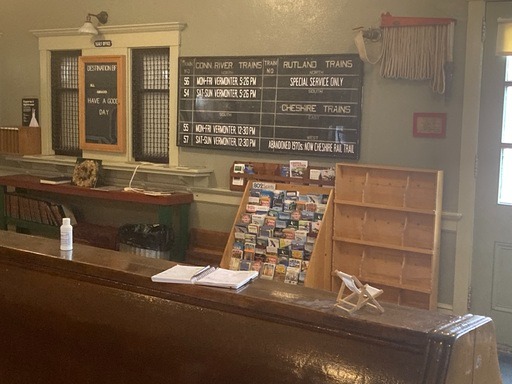 Vintage train station waiting area with schedules on a board, brochures on a rack, and a wooden bench in the foreground. Hand sanitizer sits on the counter.
