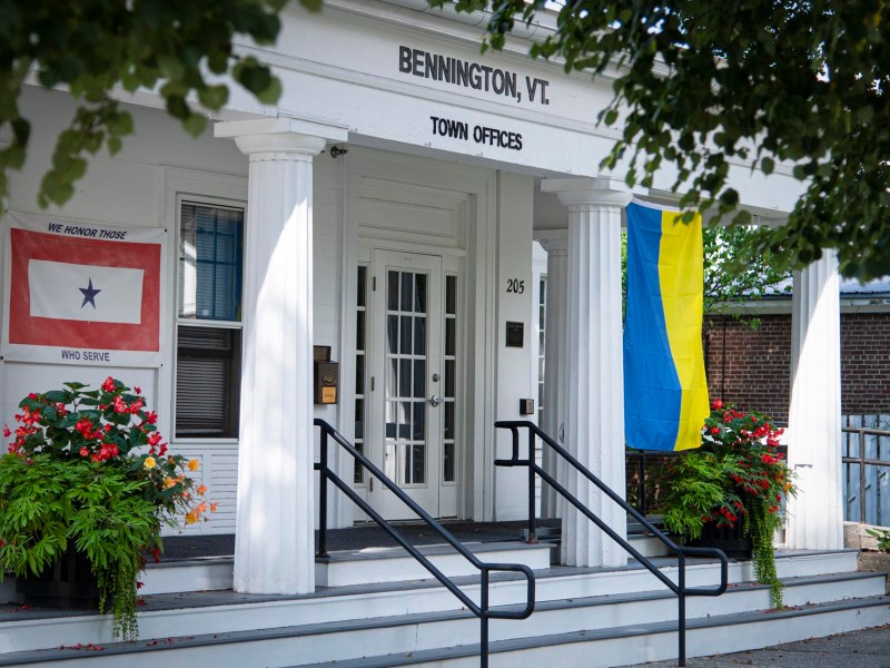 Front entrance of Bennington, VT Town Offices with a military service flag and a blue and yellow flag displayed, surrounded by potted plants and white columns.