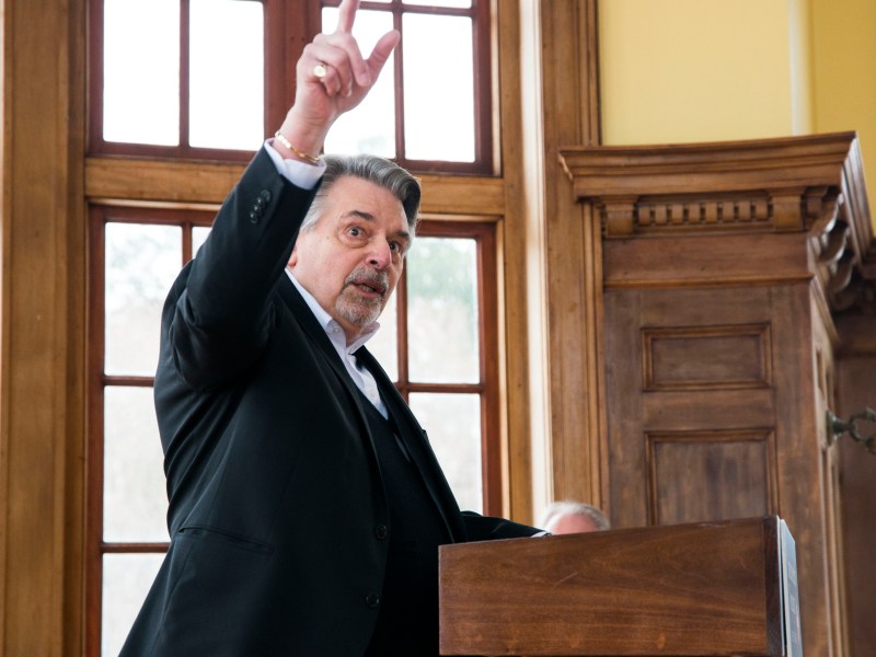 A man in a suit stands at a podium, raising one hand and speaking in a room with large windows and wooden paneling.