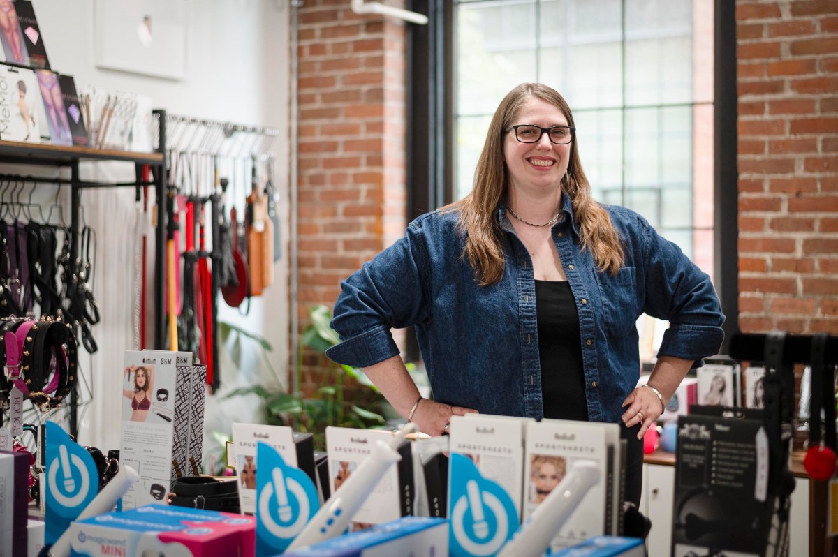 A woman stands smiling behind a display of adult products in a store with brick walls, shelves, and large windows in the background.
