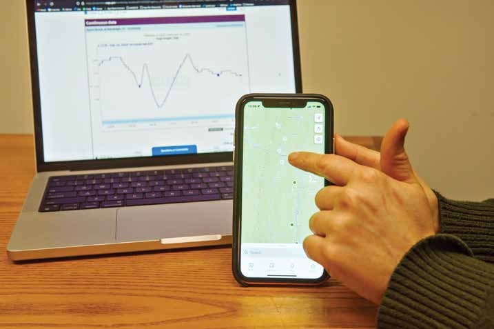Person pointing at a smartphone displaying a map, with a laptop showing a graph in the background on a wooden table.
