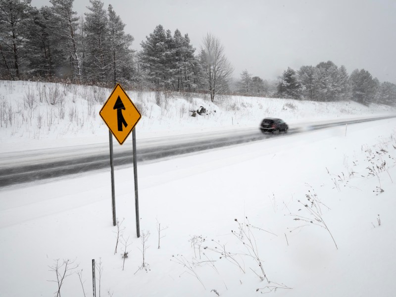 Car drives on a snow-covered road past a yellow merge sign. Snow blankets the landscape and trees, creating a wintry scene.