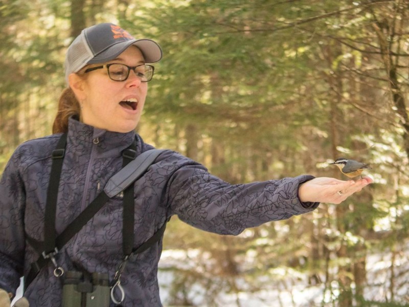 A woman stands in a forest, holding out her hand as a small bird perches on her fingers. She appears to be speaking or reacting to the bird.