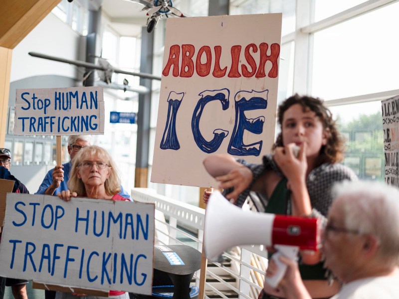 Protesters hold signs reading "Stop Human Trafficking" and "Abolish ICE" during a demonstration indoors; one person speaks into a megaphone.