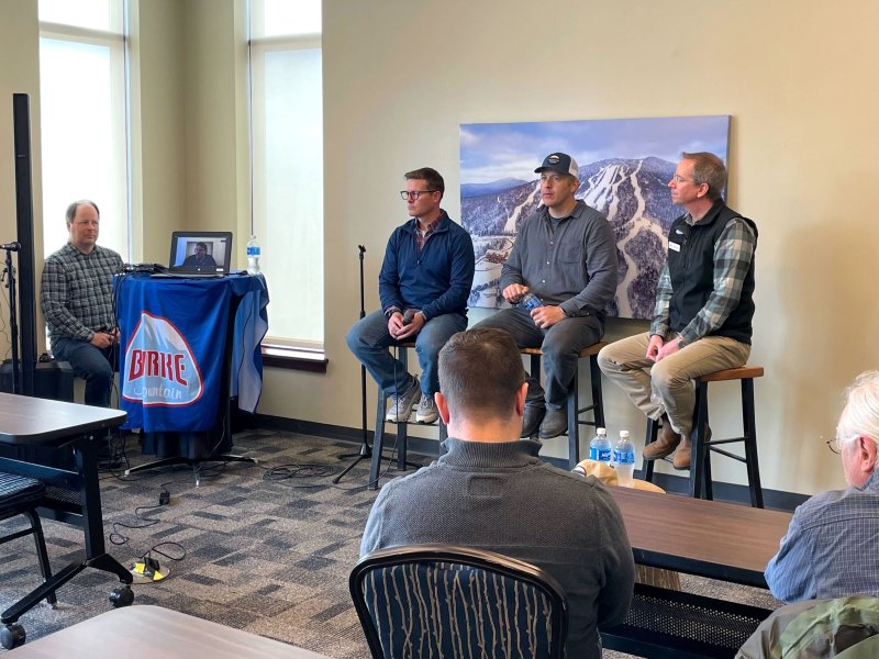 Four men are seated at the front of a conference room, three on stools and one at a table with audio equipment. Several people sit facing them. A mountain photo hangs on the wall behind them.