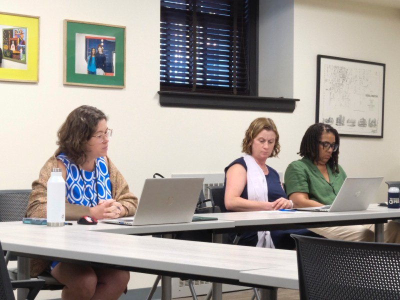 Three women sit at a conference table using laptops during a meeting in a room with framed pictures and a map on the wall.