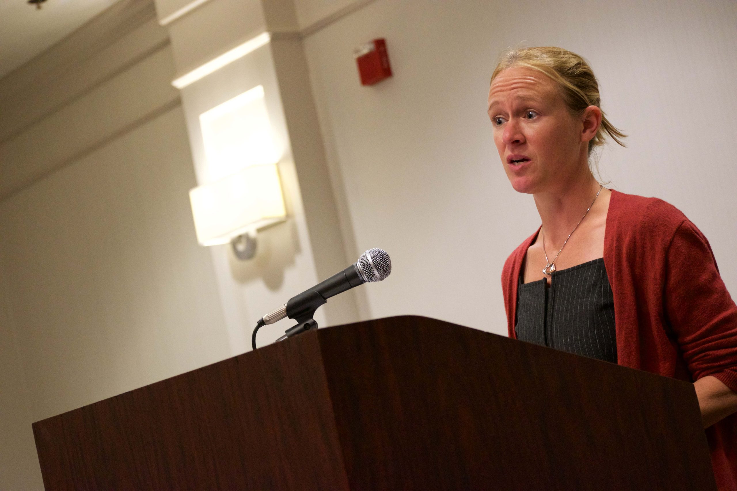 A woman stands behind a podium speaking into a microphone in a well-lit indoor setting.