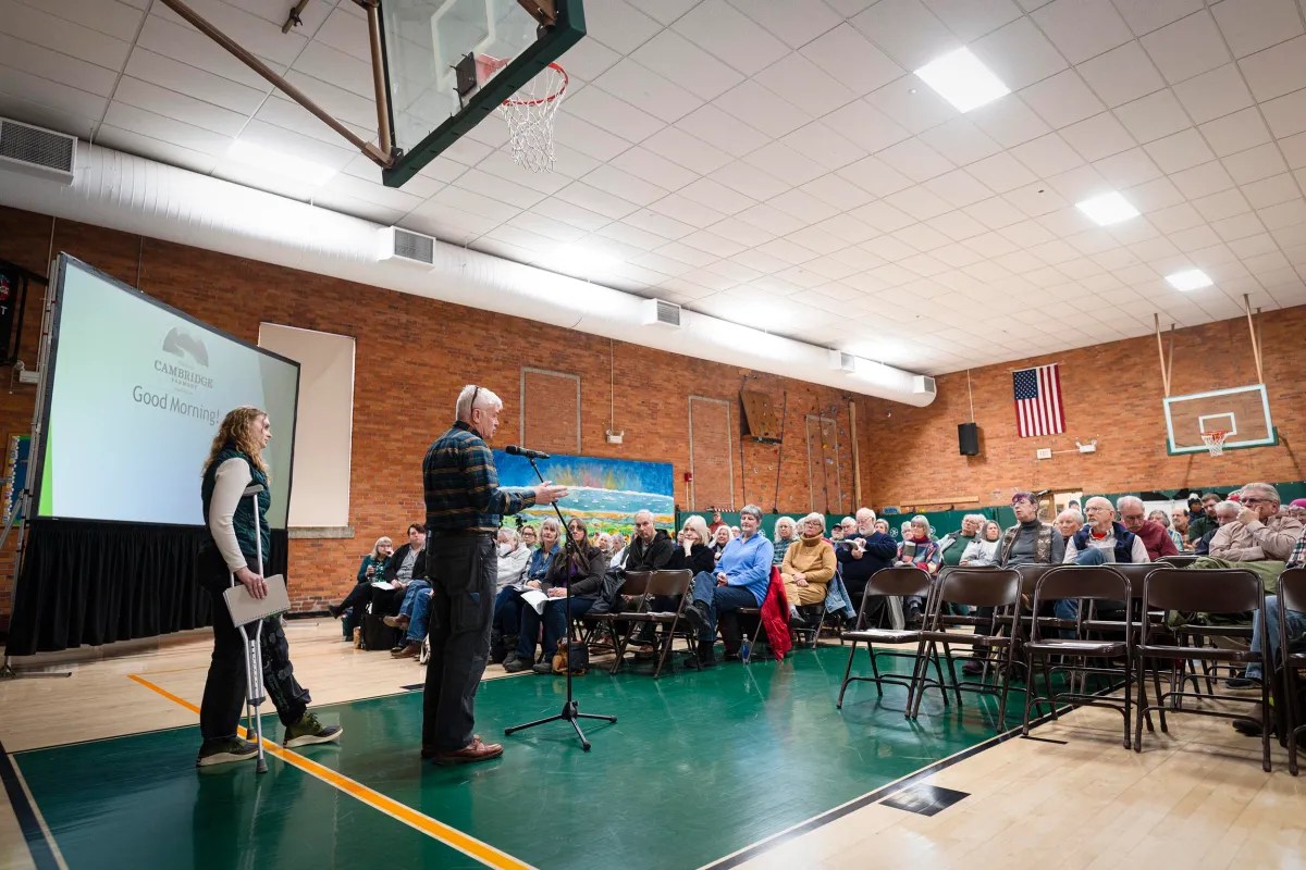 A large group of people seated in a gymnasium attending a presentation. Two individuals stand near a screen that reads "Good Morning" with a microphone on the floor. An American flag is visible.