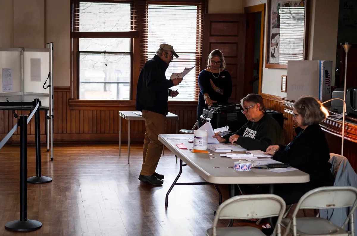People in a room checking and processing papers at a table, with one person standing and reading in a casual setting.