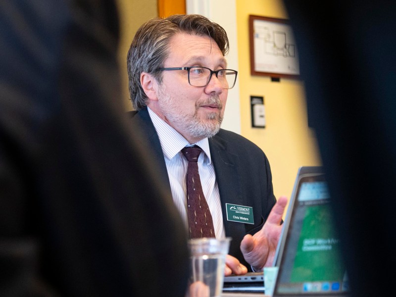 A man wearing glasses and a suit speaks during a meeting, seated at a table with laptops and a glass of water in front of him.
