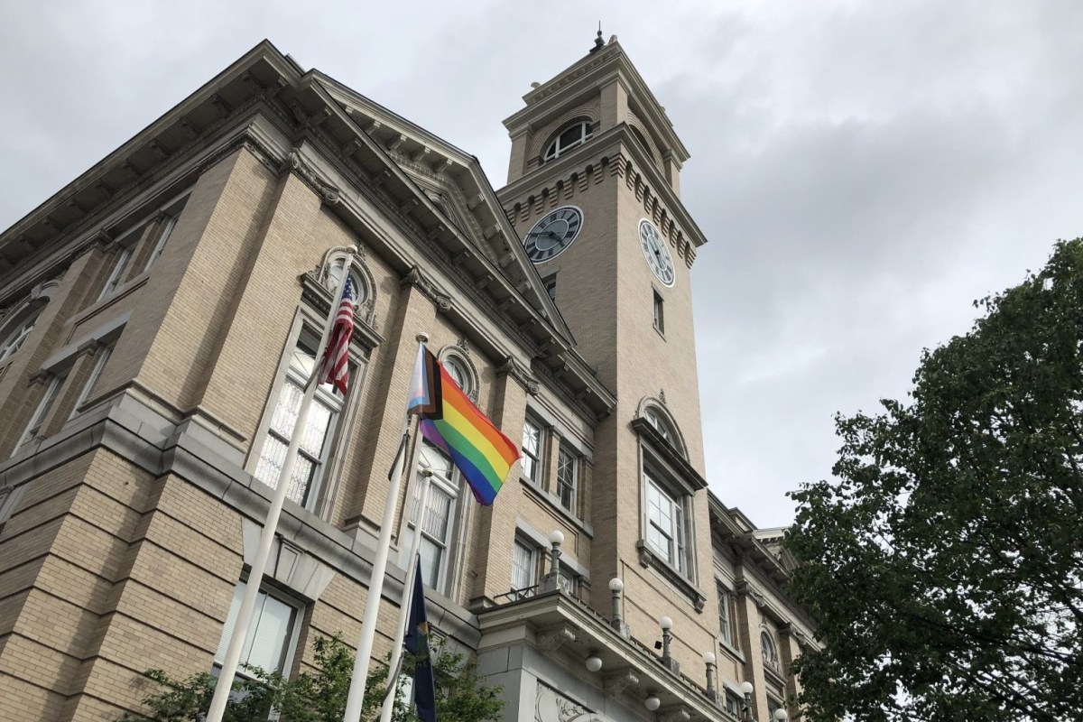 A historic building with a clock tower features two flagpoles. One flag is an American flag and the other is a rainbow pride flag. A tree is visible to the right.