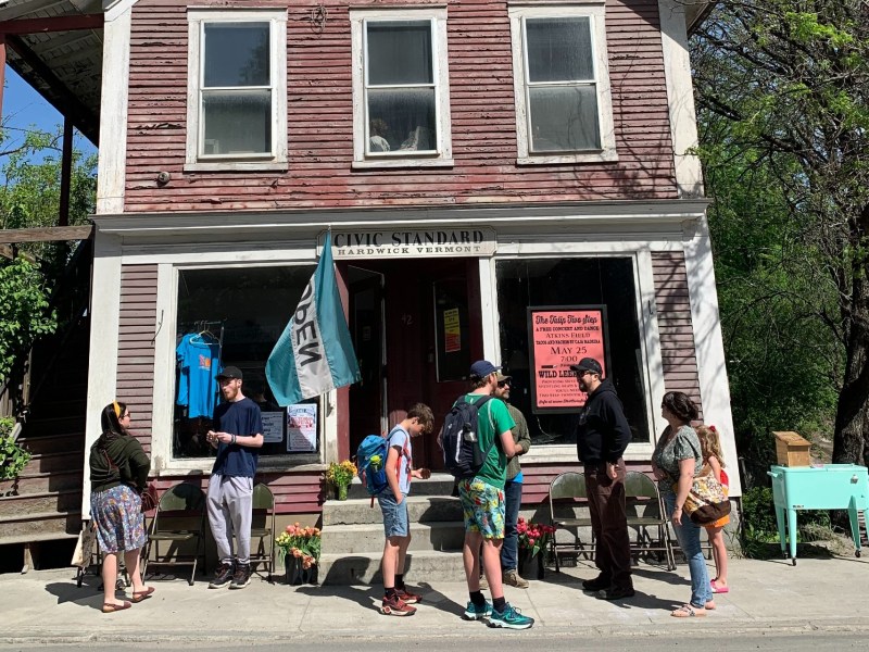 People stand outside a rustic two-story building with a sign reading "Civic Standard Hardwick Vermont." A blue open flag is displayed, and various items and plants are arranged in the front.