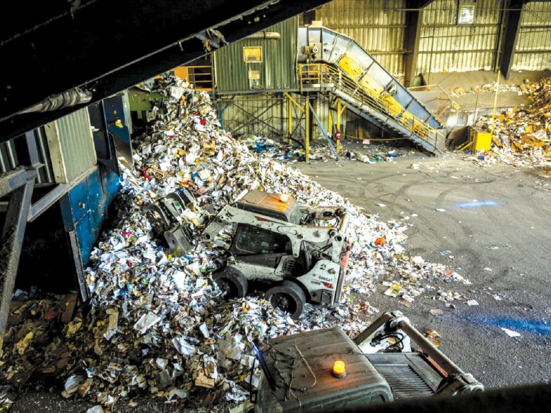 Indoor recycling facility with two loaders managing large piles of paper waste. A conveyor belt is visible in the background.