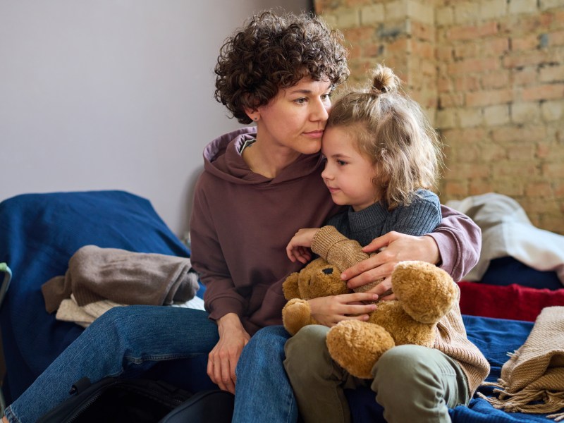 An adult and a child sit closely together on a cot; the child holds a teddy bear while the adult offers comfort in a sparse room.