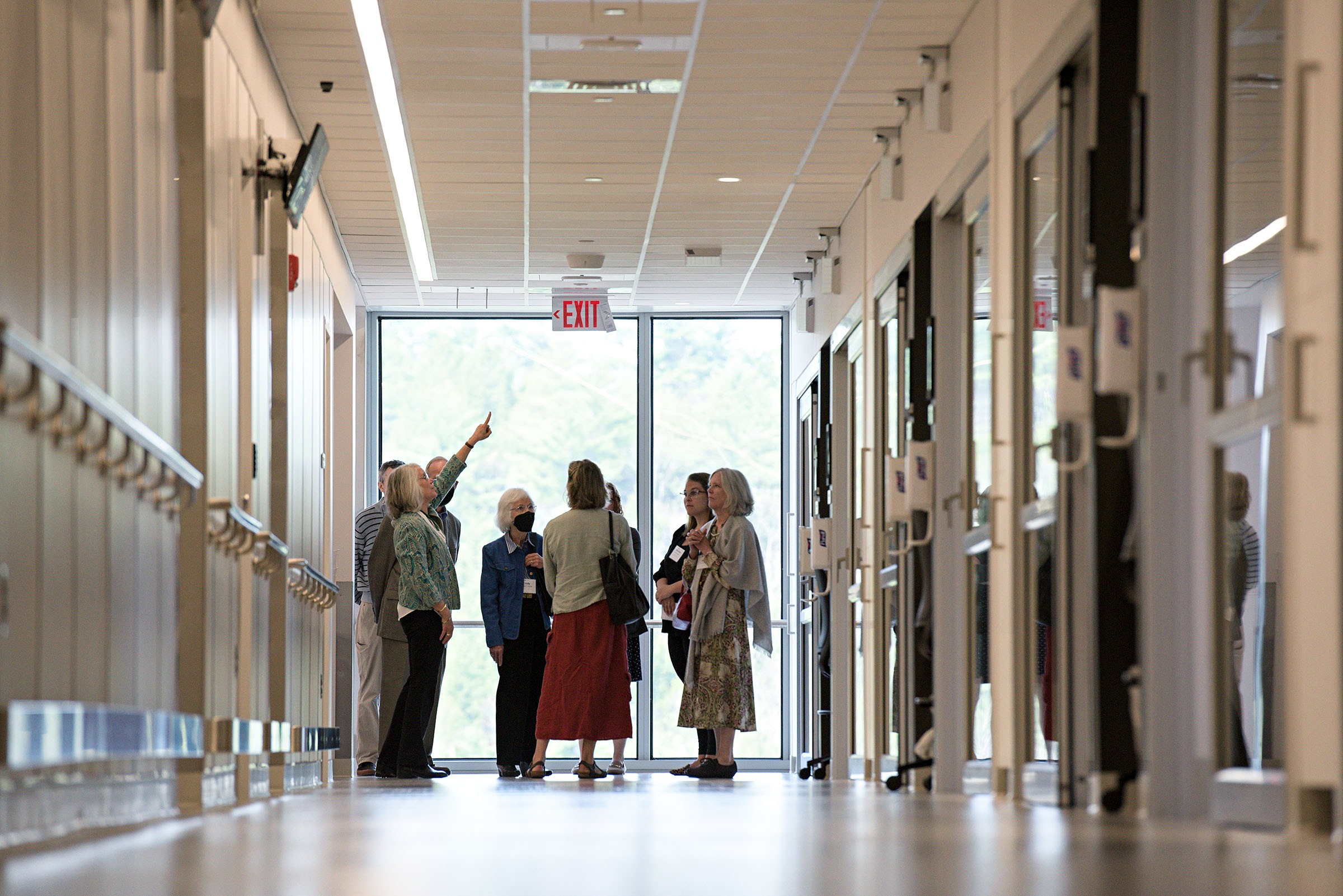A group of six adults stands in a hallway, with one person pointing upward while others look on. The hallway has large windows at the end and doors on both sides.