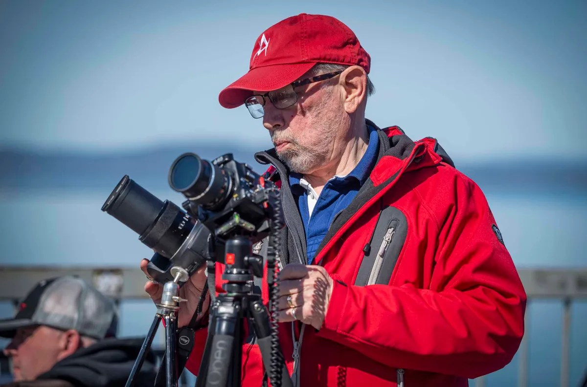 A person in a red jacket and cap operating a camera on a tripod outdoors.