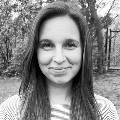 A woman with long straight hair, smiles at the camera outdoors, standing in front of trees and a fence.