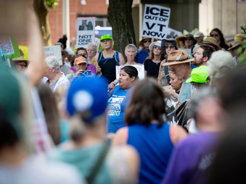 A crowd gathers at an outdoor protest holding signs, including ones that read “ICE OUT OF VT NOW” and “NOT ONE MORE.” A person speaks into a microphone at the center.
