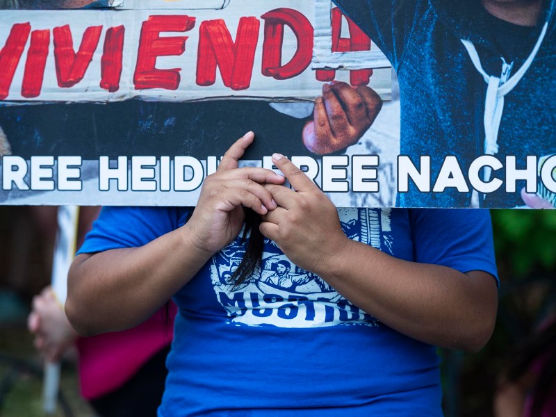 A person holds a banner that reads "FREE HEIDI FREE NACHO!" and part of the word "VIVIENDA," with their hands visible in front of their torso.