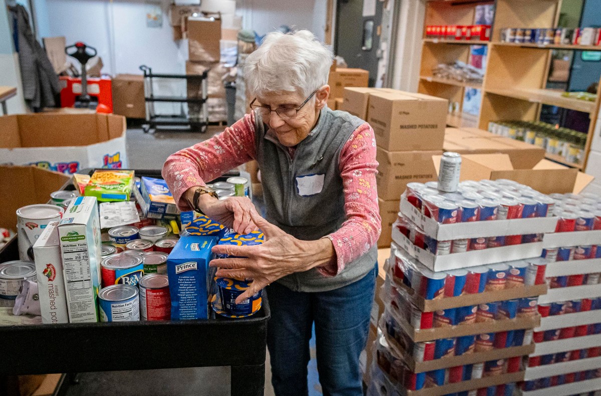 A woman organizes canned goods on a cart in a food pantry, surrounded by shelves and stacks of boxes.