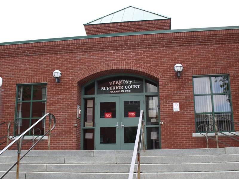 Entrance of a red brick building with a sign above the door reading "Vermont Superior Court Franklin Unit"; concrete steps lead up to double glass doors.