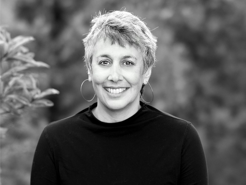 A person with short, gray hair and hoop earrings smiles at the camera, wearing a black top, with a blurred natural background.