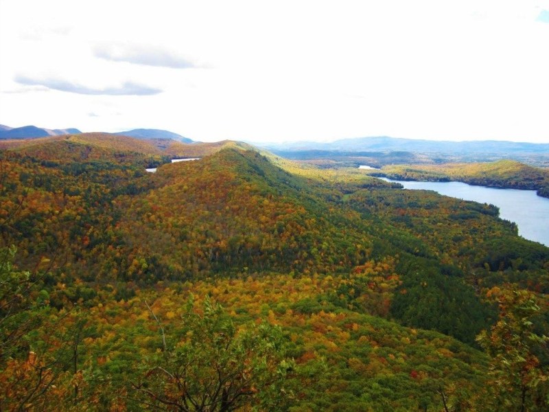 A scenic view of rolling hills covered in autumn foliage with a river or lake winding through the landscape under a partly cloudy sky.