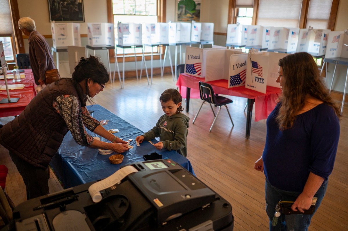 A child receives a voting sticker from a woman at a polling station, while another woman stands nearby. Voting booths and a ballot machine are visible in the background.