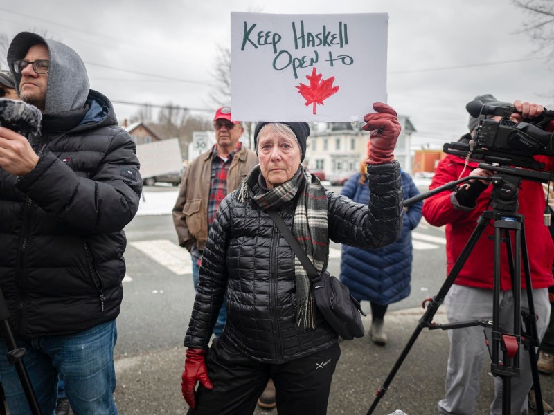 A person holds a sign reading "Keep Haskell Open" with a red maple leaf. They stand among a group of people, some holding cameras, outdoors on an overcast day.