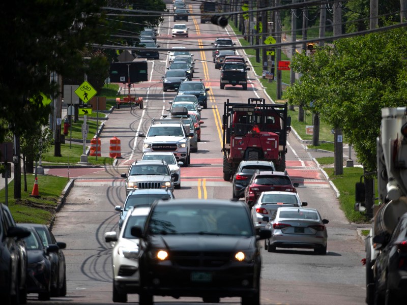 A busy two-lane street with multiple cars and a truck driving in both directions, surrounded by trees and traffic signs on a sunny day.