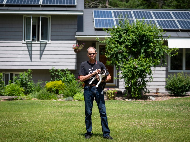 A man stands on a lawn holding a dog in front of a house with solar panels on the roof.