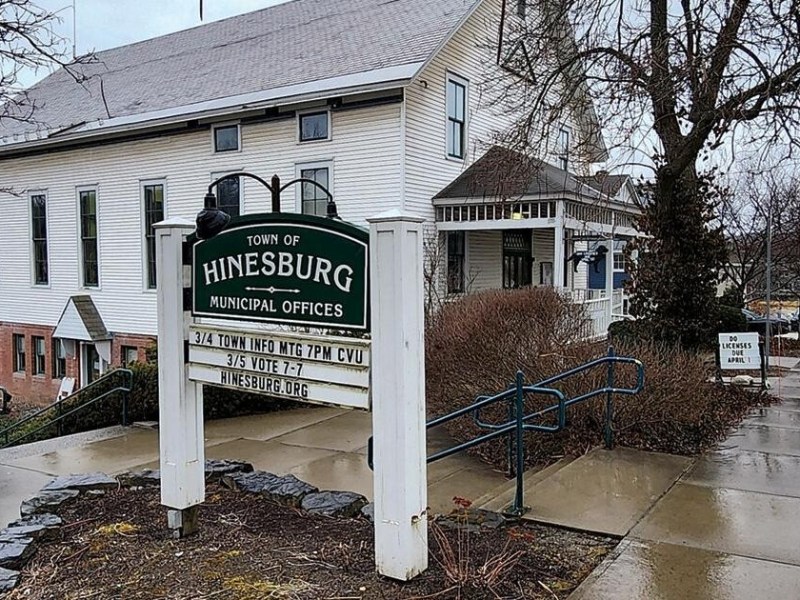 Hinesburg Municipal Offices building with a sign listing town meeting information, surrounded by wet pavement and bare trees on a cloudy day.