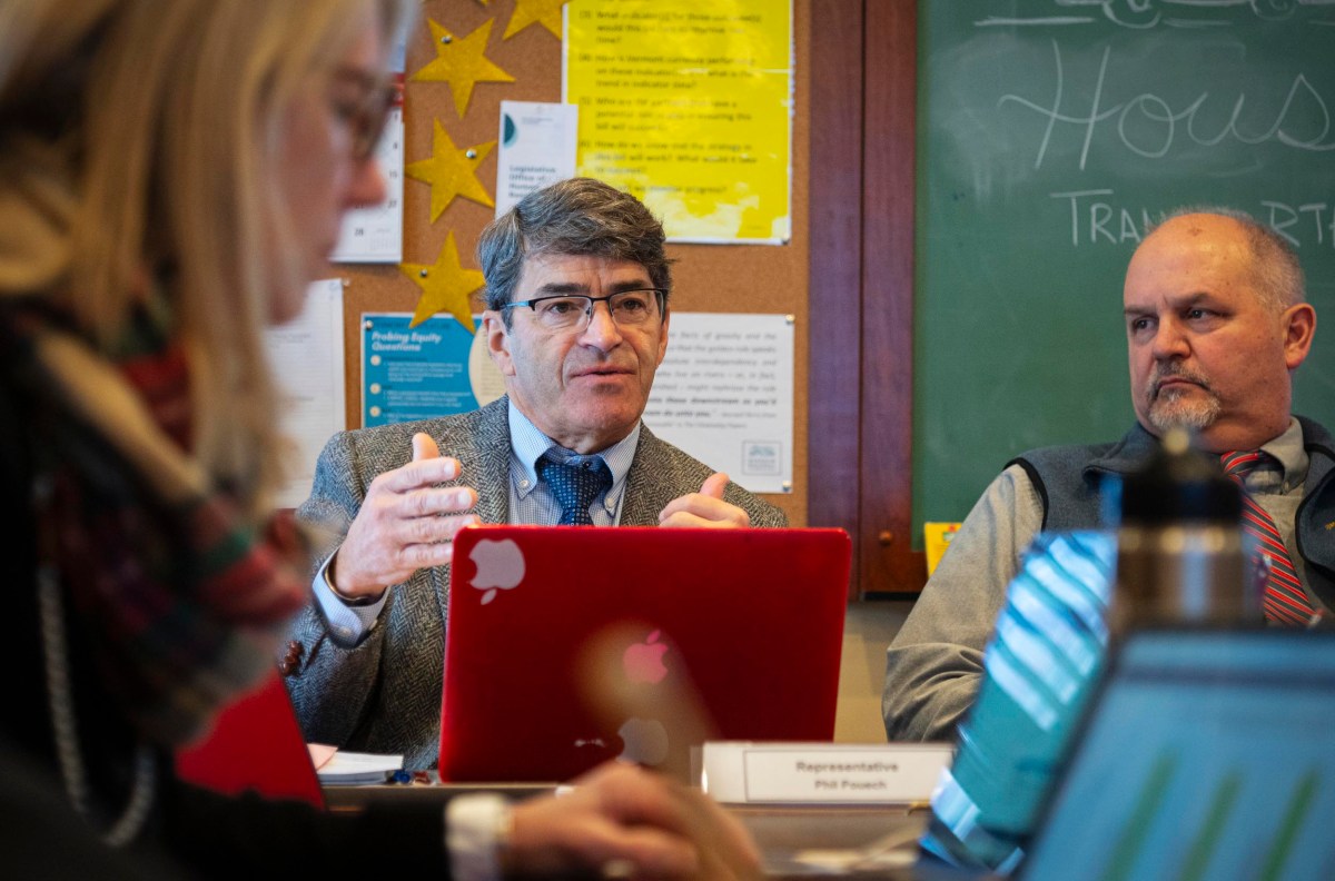 A group of people engaged in a discussion around a table with laptops and notes. A man in the center gestures while speaking, with a red laptop in front of him. Bulletins and chalkboard in the background.