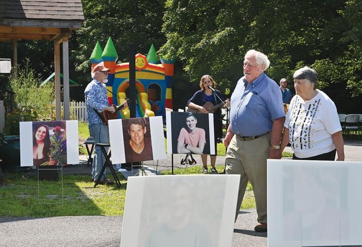 Two people walk past large portrait photos on display at an outdoor event with musicians performing and a colorful bounce house in the background.