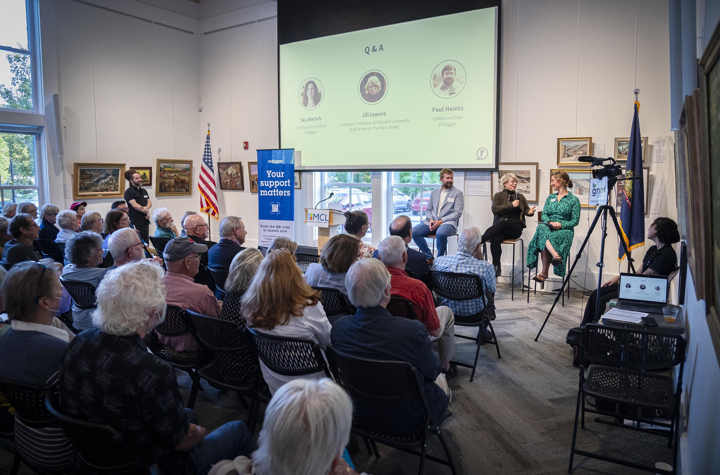 A panel discussion in progress with three speakers on stage and an audience seated in a well-lit room, a camera filming the event, and a screen displaying Q&A and images of the speakers.