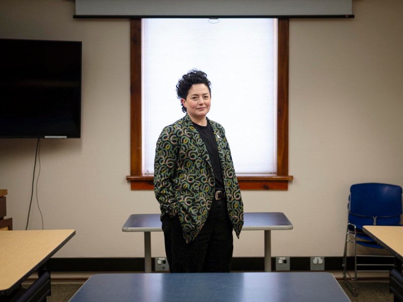 Person standing in a classroom between desks, wearing a patterned jacket with hands in pockets.