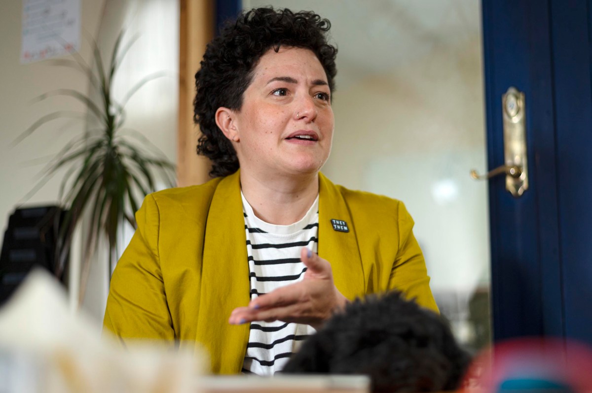 A person wearing a yellow blazer and striped shirt gestures while speaking indoors, with a plant and door in the background.