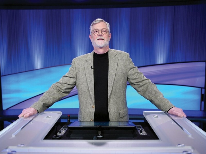 A man with gray hair and beard, wearing glasses, a gray blazer, and a black shirt, stands behind a game show podium on a blue-lit stage.