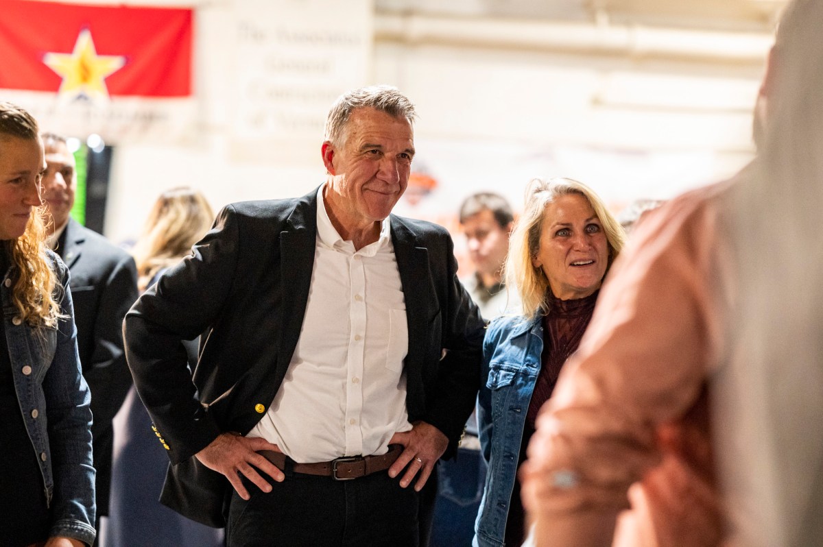A man in a black blazer and a woman in a denim jacket are standing in a crowd, with a red flag hanging in the background.