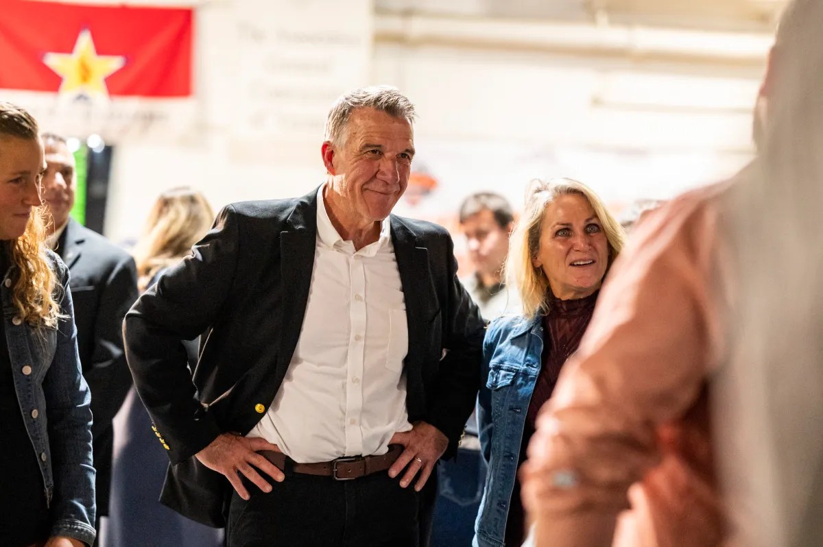 A man in a black blazer and a woman in a denim jacket are standing in a crowd, with a red flag hanging in the background.