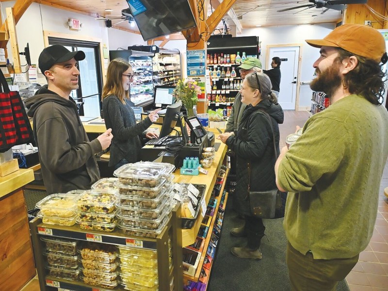 People stand and chat at a store checkout counter; one person checks out while others wait, surrounded by packaged food and drinks.