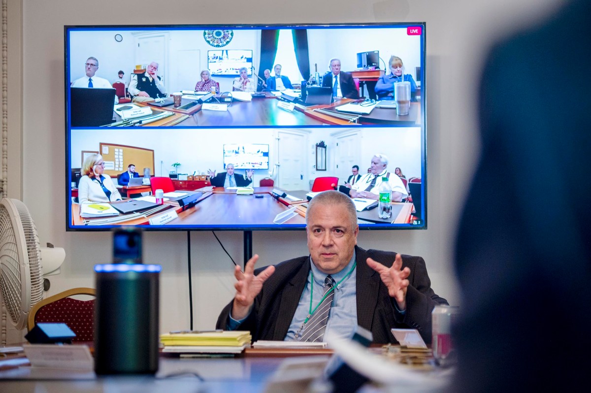 A man sits at a conference table speaking, with a large screen behind him showing multiple people in a video meeting.