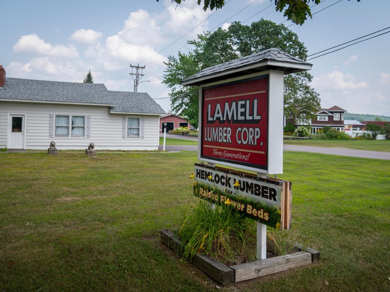 A large outdoor sign reads "Lamell Lumber Corp." and "Hemlock Lumber Raised Flower Beds," standing on a grassy lawn beside a white house.