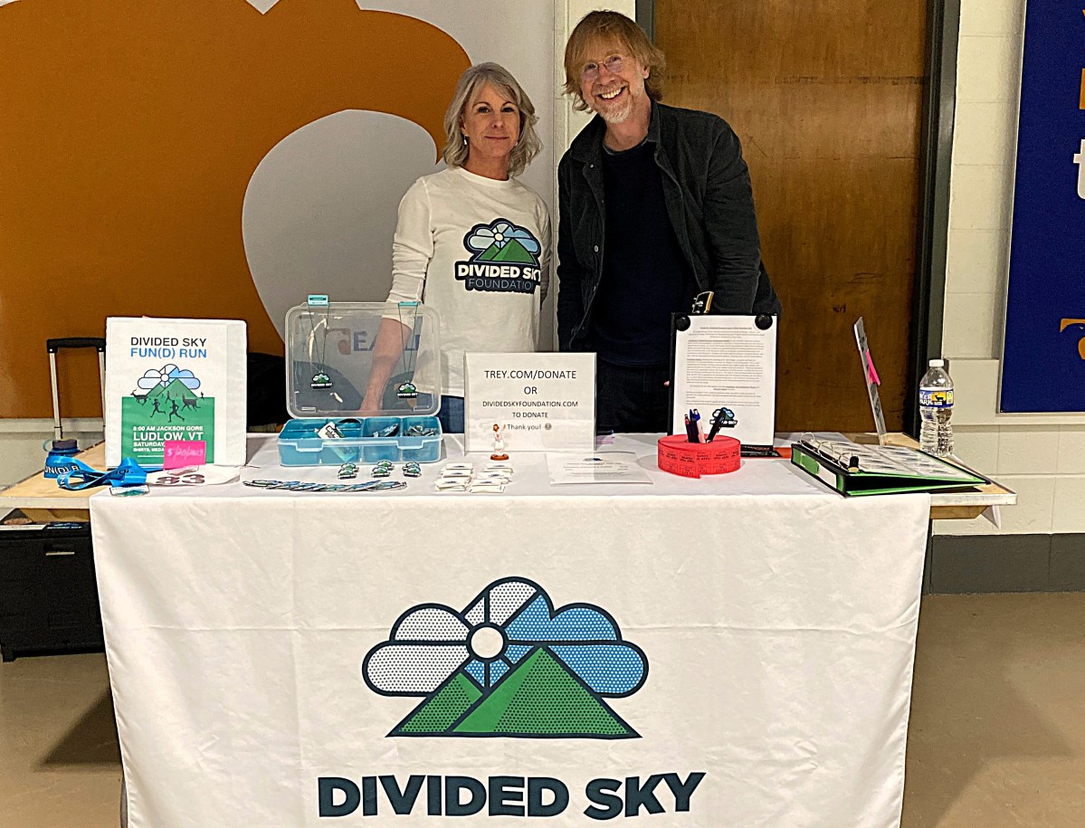 Two people stand behind a table with a "Divided Sky" banner, promotional materials, and informational signs displayed in a room with white walls and a wooden door.
