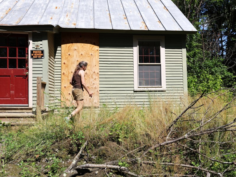 A person walks towards a small house with a boarded-up window, a red door, and overgrown grass in the front yard.